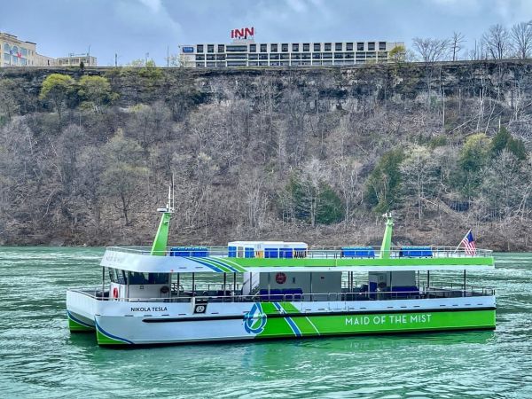 The Maid of the Mist floats on a river with trees and a hotel in the background.