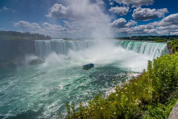 The Maid of the Mist in Horseshoe Falls under a partly cloudy sky, surrounded by lush greenery.