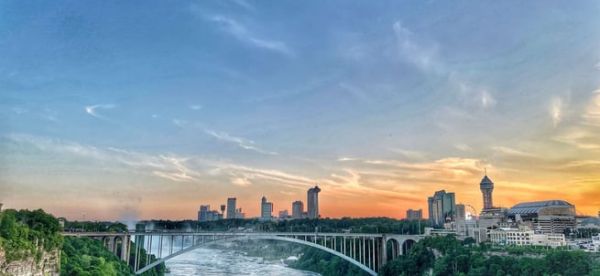 A scenic view of a bridge over a river at sunset, with the Niagara Falls skyline and lush greenery.