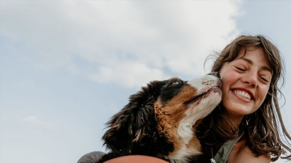 一张可爱的棕发女孩自拍：她身着运动装，蹲着身子，微笑着抱着一个球，而她那只三色毛茸茸的狗狗正亲吻她的脸颊。闭上眼睛，感受你的爱